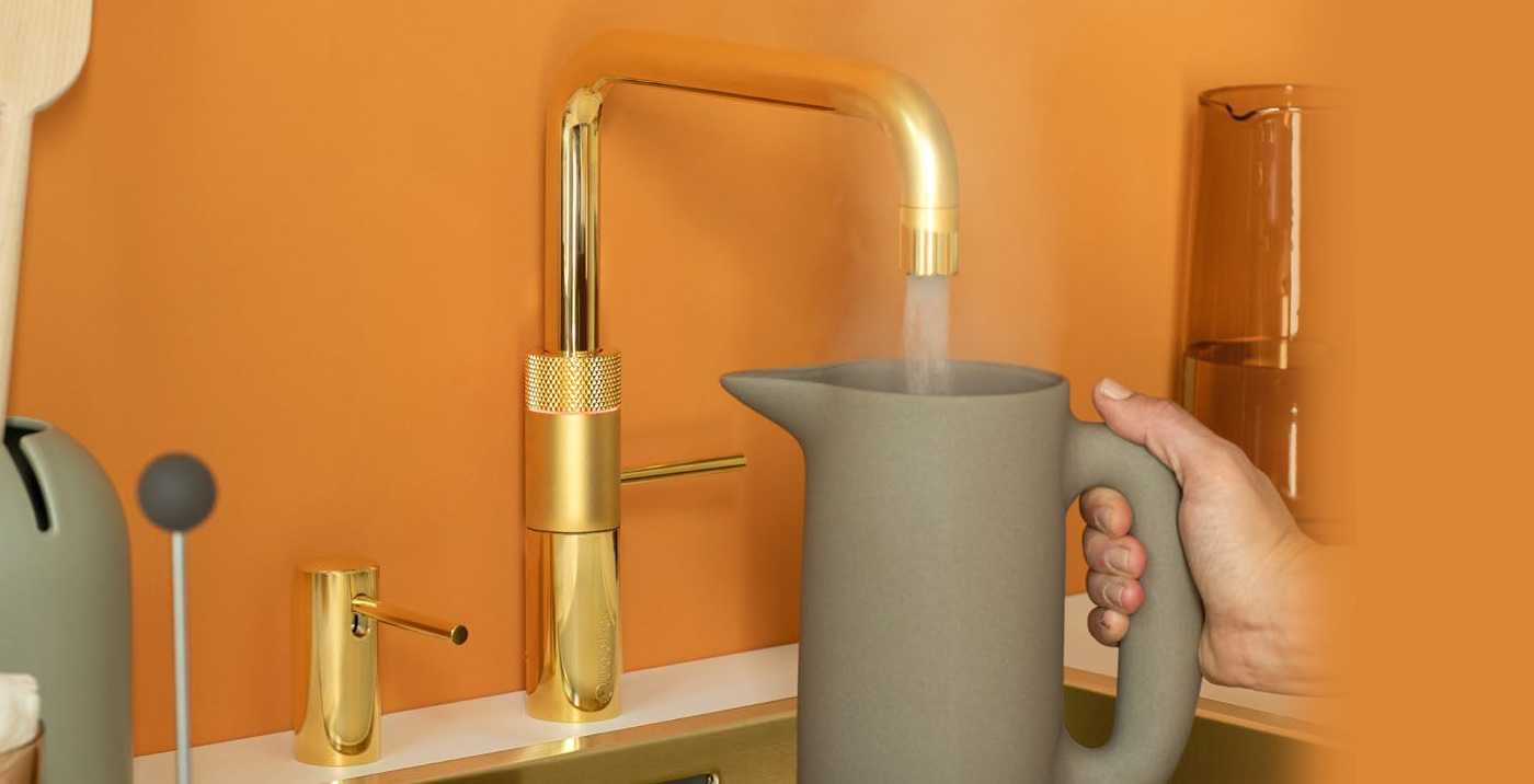 A gold Quooker Fusion Square tap pouring boiling water into a stoneware jug against an orange wall